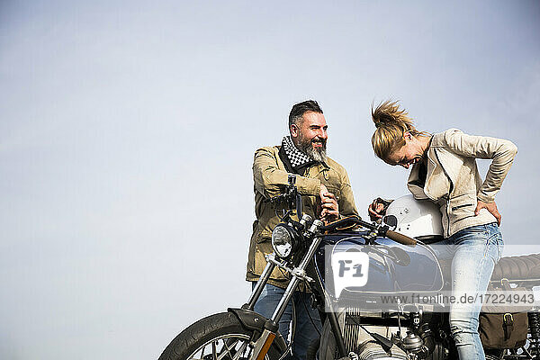 Smiling man talking with cheerful female biker sitting on motorcycle by sky