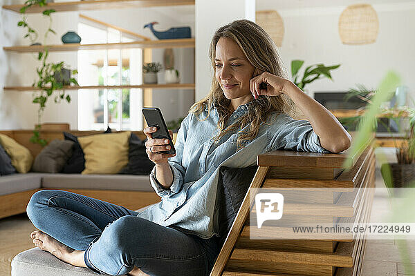 Blondhaarige Frau  die im Wohnzimmer sitzend eine SMS über ihr Smartphone verschickt