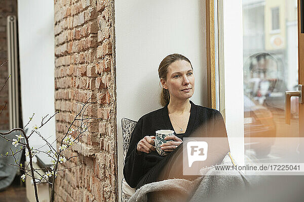 Nachdenkliche Frau mit Teetasse  die zu Hause am Fenster sitzt