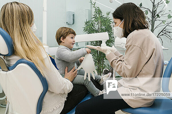 Kinderzahnarzt mit Gesichtsschutzmaske im Gespräch mit einem Jungen  der mit seiner Mutter in der Klinik sitzt