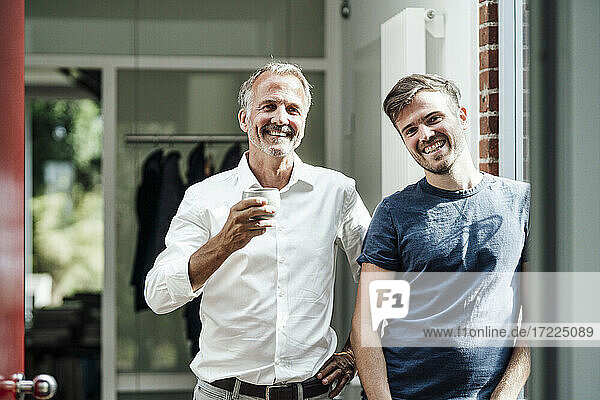 Smiling father with coffee cup standing by son leaning on wall during sunny day
