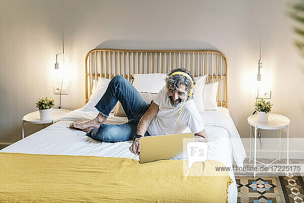 Entspannter älterer Mann mit Kopfhörern und Laptop auf dem Bett zu Hause