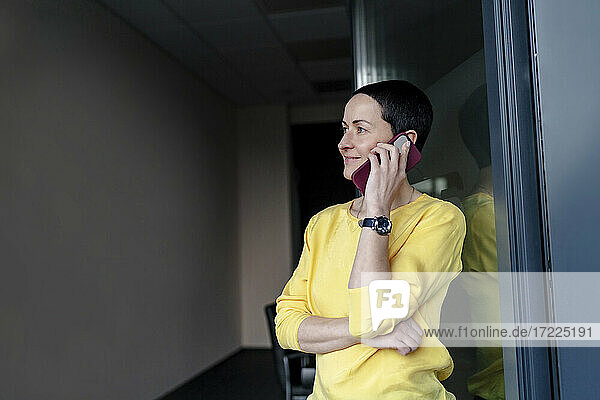 Lächelnde Geschäftsfrau  die über ihr Smartphone in einem modernen Büro nachdenkt