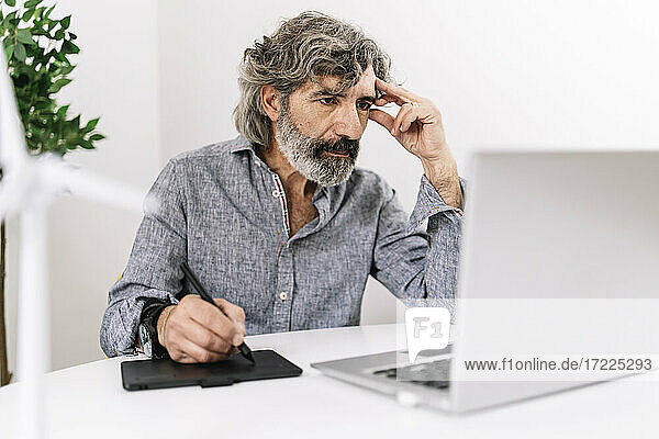 Konzentrierter älterer Geschäftsmann mit Grafiktablett am Laptop  während er im Büro sitzt