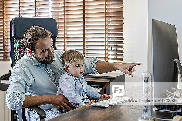 Lächelnder Vater  der auf einen Computer zeigt  während sein Sohn zu Hause auf dem Schoß sitzt