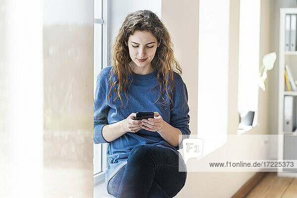Woman with brown hair texting message through smart phone while sitting at window in living room