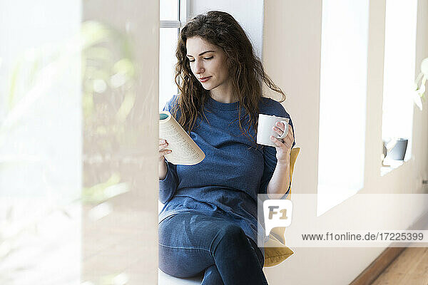 Schöne Frau liest ein Buch  während sie einen Kaffee am Fenster im Wohnzimmer trinkt