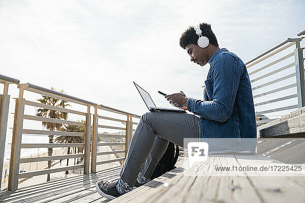 Afro man using smart phone over laptop while sitting on steps at promenade