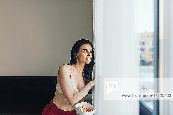 Mature woman holding bowl of fruit while looking through window at home