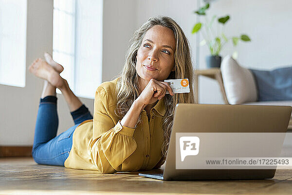 Nachdenkliche Frau mit Kryptowährungs-Kreditkarte  die wegschaut  während sie zu Hause vor ihrem Laptop liegt