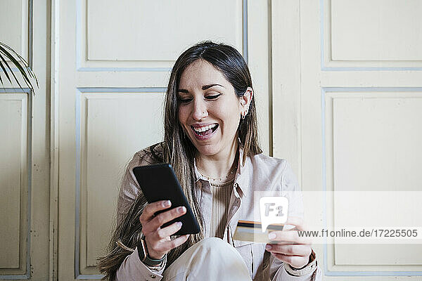 Happy woman using smart phone while holding credit card at home