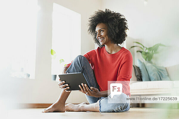Lächelnde Frau hält digitales Tablet  während sie zu Hause auf dem Boden sitzt