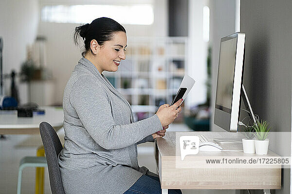 Lächelnde Unternehmerin  die ein Mobiltelefon benutzt und ein Dokument im Büro zu Hause hält