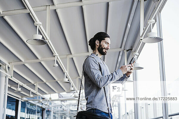 Lächelnder Geschäftsmann mit Laptoptasche  der ein Mobiltelefon auf dem Balkon eines Büros benutzt