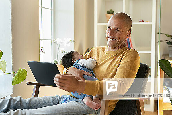 Glücklicher Vater hält schlafendes Baby und digitales Tablet  während er zu Hause wegschaut