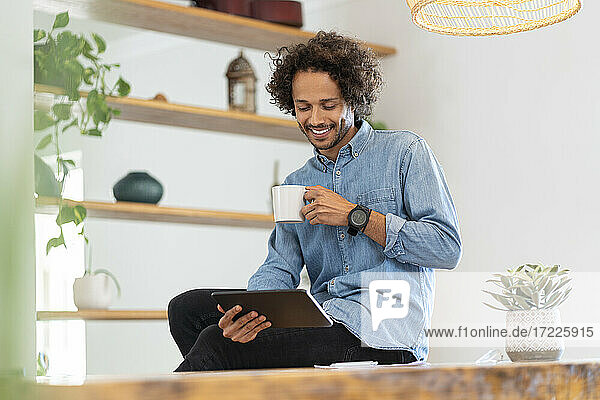 Lächelnder männlicher Fachmann  der ein digitales Tablet benutzt und eine Kaffeetasse zu Hause hält