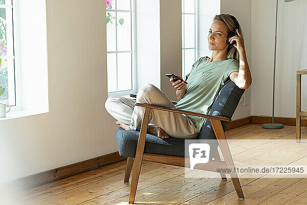 Nachdenkliche Frau hört Musik und hält ein Smartphone auf einem Stuhl zu Hause