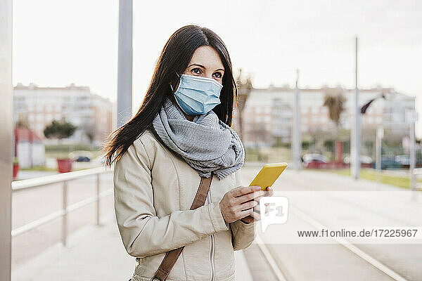 Mid adult woman with protective face mask looking away while holding mobile phone at railroad station
