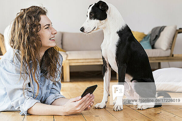 Lächelnde Frau mit Mobiltelefon  die einen Jack Russell Terrier betrachtet  während sie im Wohnzimmer auf dem Boden liegt