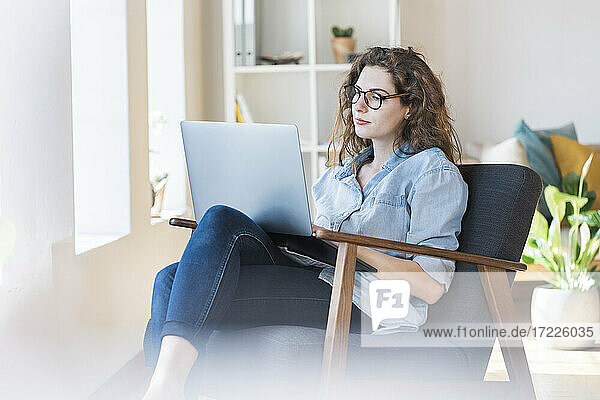 Frau  die einen Laptop benutzt und auf einem Sessel im Wohnzimmer sitzt