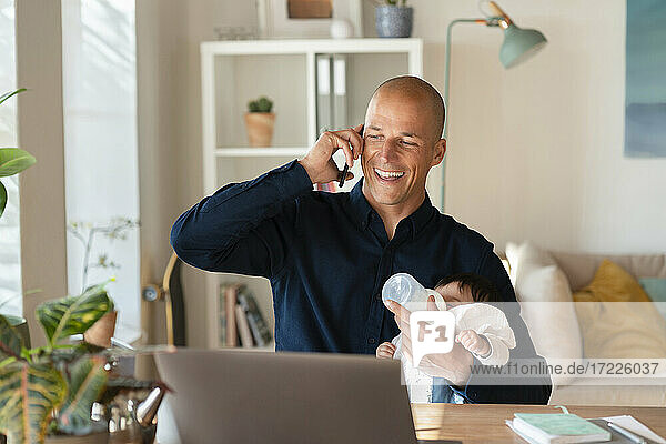 Smiling father talking on smart phone while feeding milk to daughter at home