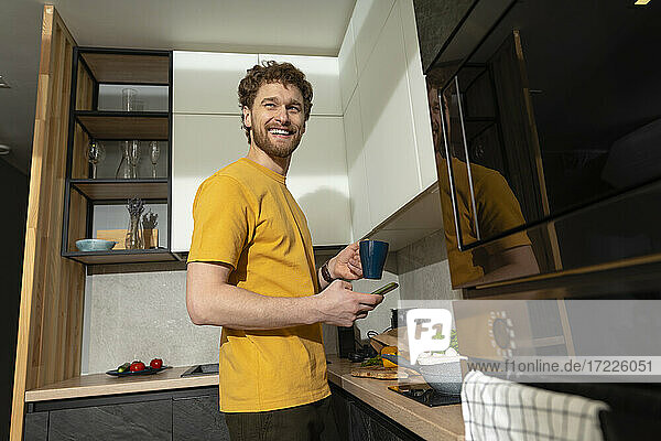 Lächelnder Mann  der wegschaut  während er ein Smartphone und ein digitales Tablet in der Küche hält