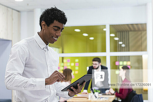 Lächelnder Geschäftsmann  der ein digitales Tablet benutzt  mit Kollegen im Hintergrund im Büro