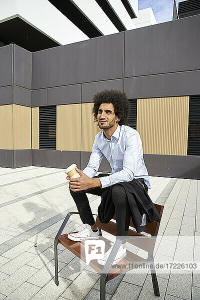 Man holding coffee cup while sitting on chair at street in city