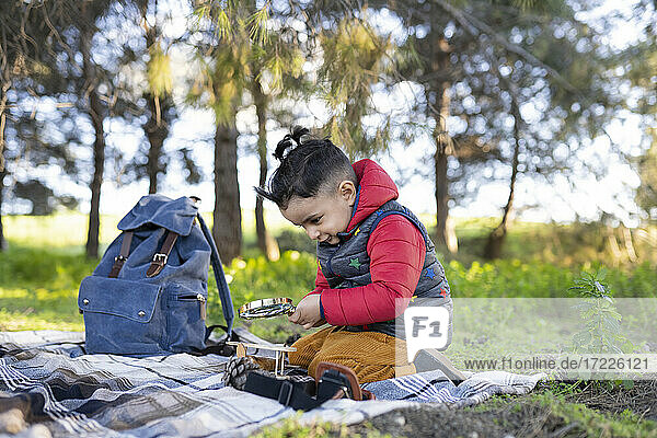 Neugieriger Junge  der auf einer Picknickdecke kniend ein Vergrößerungsglas durchsucht