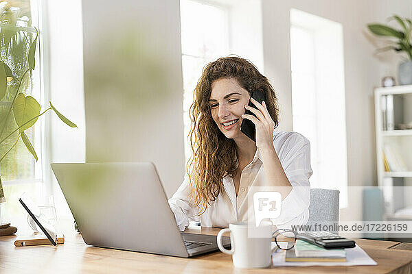 Zufriedene Unternehmerin  die mit einem Smartphone telefoniert  während sie einen Laptop am Schreibtisch im Heimbüro benutzt