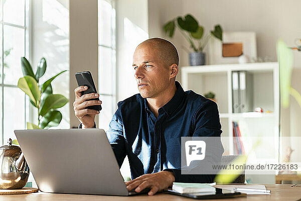 Männlicher Unternehmer arbeitet am Smartphone  während er zu Hause am Laptop sitzt