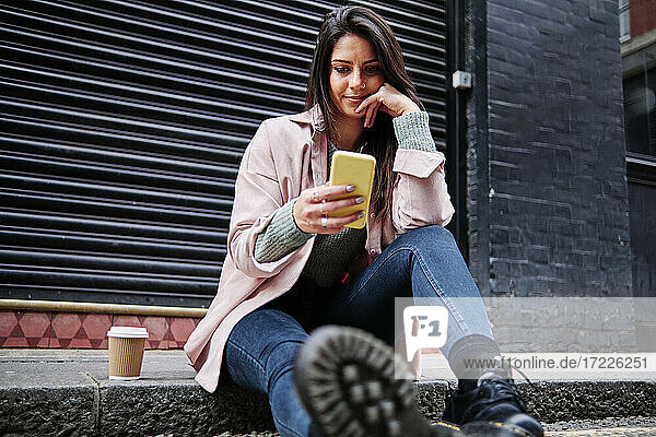 Schöne Frau mit Kaffeetasse und Mobiltelefon auf dem Bürgersteig sitzend