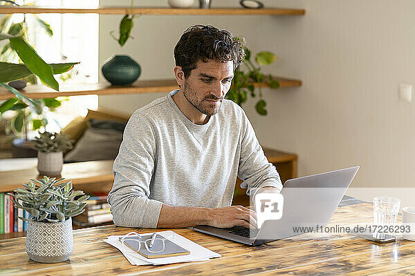 Konzentrierter männlicher Freiberufler sitzt am Tisch und arbeitet von zu Hause aus am Laptop