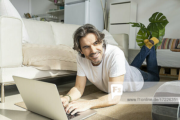 Thoughtful man with laptop looking away while lying on floor at home