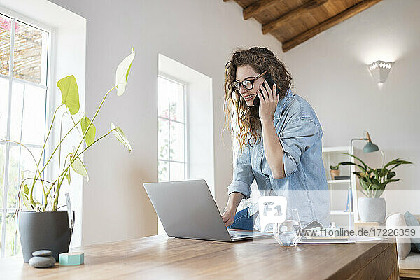 Lächelnde weibliche Fachkraft  die mit einem Smartphone telefoniert  während sie an einem Laptop am Schreibtisch arbeitet