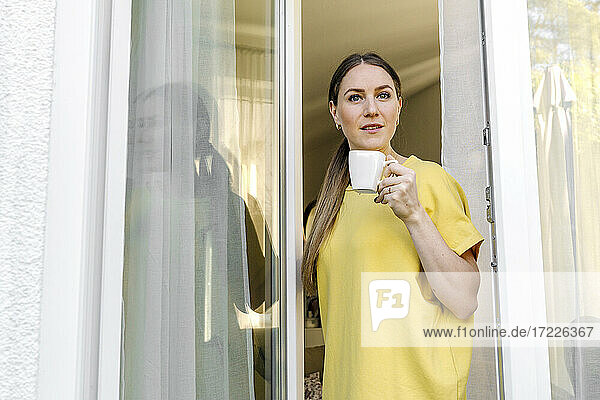 Braunhaarige Frau mit Kaffeetasse am Fenster stehend