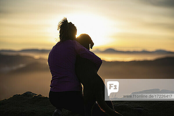 Junge Frau umarmt ihren Hund bei Sonnenaufgang
