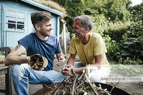 Son holding firewood discussing with father while crouching in backyard