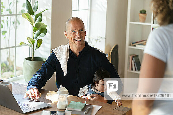 Lächelnder Mann sitzt mit Baby und sieht Frau bei der Arbeit zu Hause an