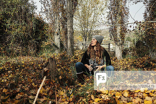 Woman with camera looking away while sitting amidst autumn leaves in forest