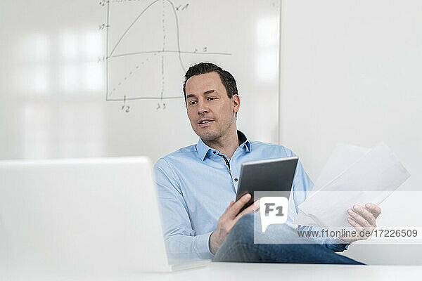 Männlicher Fachmann  der auf einen Laptop schaut  während er ein digitales Tablet und Dokumente im Büro hält