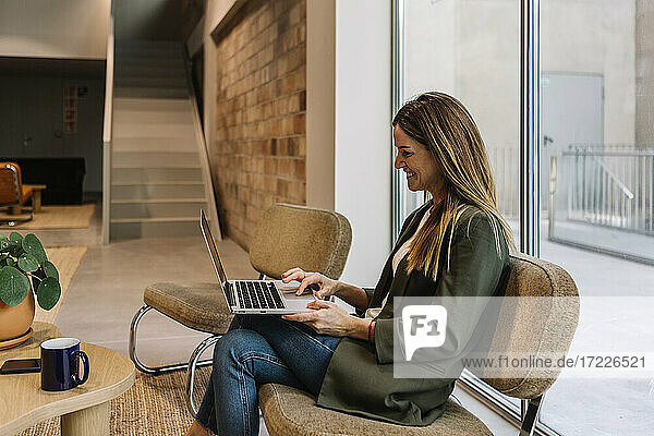 Lächelnde Geschäftsfrau  die einen Laptop benutzt und auf einem Stuhl im Büro sitzt