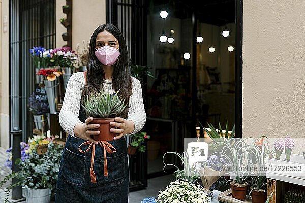 Junge Floristin mit Gesichtsschutzmaske  die eine Topfpflanze hält  während sie in einem Blumenladen arbeitet