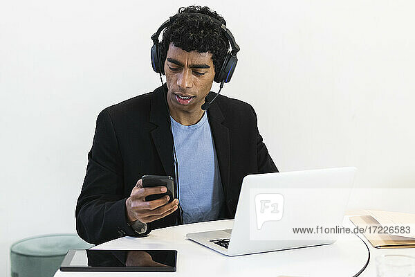 Junger Geschäftsmann mit Headset und Laptop  der ein Mobiltelefon benutzt