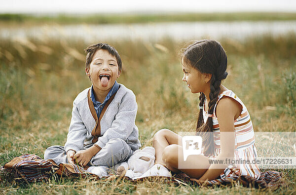 Mädchen schaut Bruder mit Down-Syndrom an  der die Zunge herausstreckt  während er im Gras sitzt