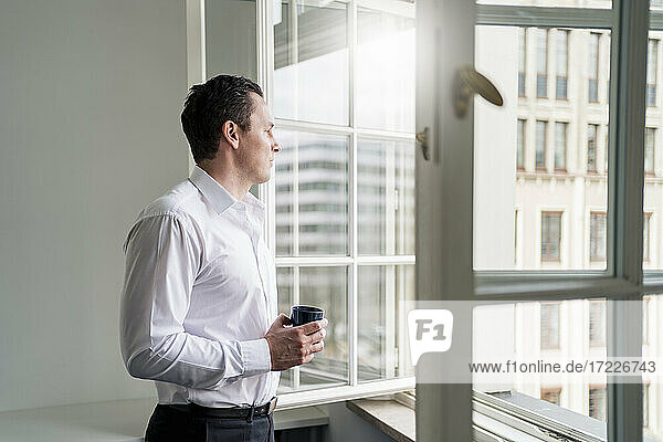 Männlicher Unternehmer  der eine Kaffeetasse hält und durch ein Fenster am Arbeitsplatz schaut