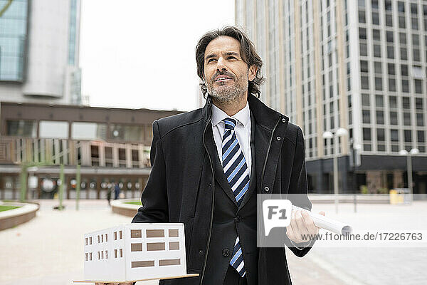 Männlicher Architekt  der wegschaut  während er ein architektonisches Modell und eine Blaupause gegen ein Bürogebäude hält