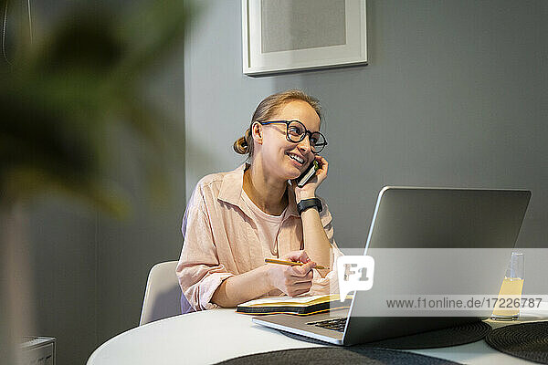Lächelnde Frau mit Laptop und Buch  die zu Hause mit ihrem Mobiltelefon spricht