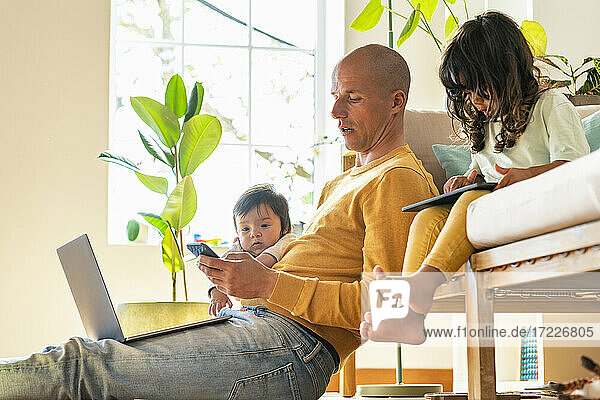 Vater benutzt Smartphone  während Baby und Kleinkind bei ihm zu Hause sitzen