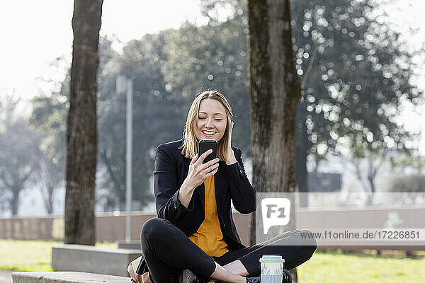 Lächelnde Geschäftsfrau  die ein Smartphone benutzt  während sie im Schneidersitz im Park sitzt